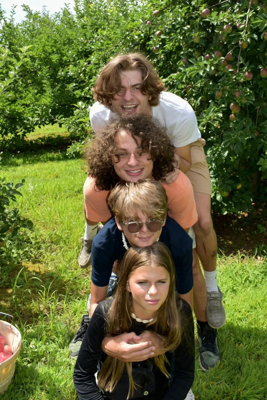 Four young people stacked in playful totem pole formation in apple orchard on sunny day